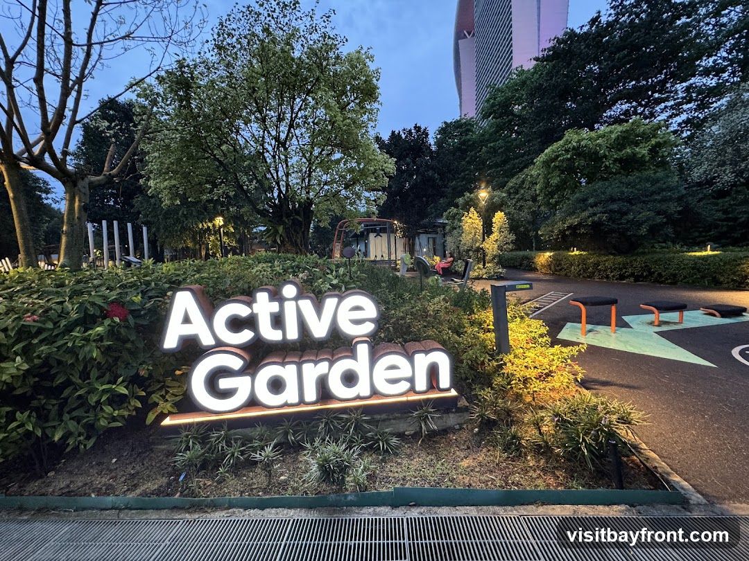 A lit sign that reads "Active Garden" is prominently displayed in a garden area. The sign is illuminated with white light and has a textured brown border. Behind the sign, lush green bushes and trees are visible. In the background, there are outdoor exercise equipment, benches, and a tall building. The scene is set in the evening or twilight.