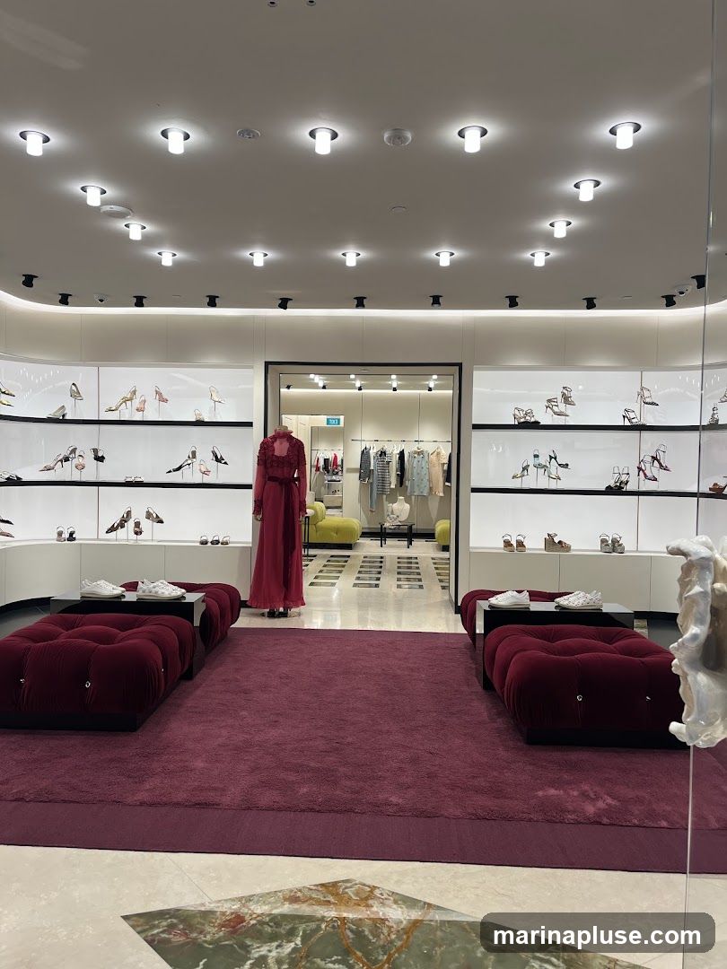 The interior of a high-end store displays a curated selection of women's footwear and apparel. Shelves along the walls are filled with various styles of high-heeled shoes and sandals. In the center, a mannequin is dressed in an elegant, long red gown. Deeper within the store, racks of clothing are visible, along with plush seating areas in lime green. Two deep red velvet benches are positioned in the foreground, each with a display of white sneakers. The store features bright, modern lighting and a luxurious ambiance.
