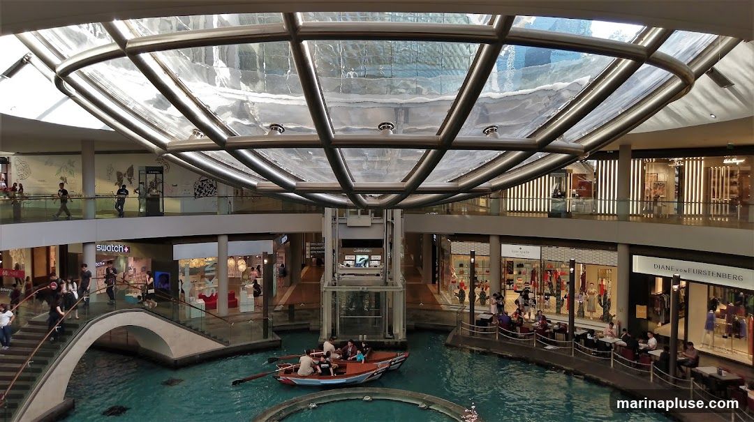 An interior view of a modern shopping mall featuring a canal with a boat. The mall has multiple levels with various shops visible, including Swatch, Chanel, Calvin Klein, Kate Spade, and Diane von Furstenberg. People are seen walking on escalators and bridges, and dining at cafes along the canal. The ceiling is a large glass structure with metal beams.
