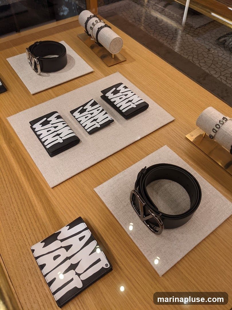 A display case shows various accessories, including black leather belts and wallets with the bold white text "WANT WANT" printed on them. There are also bracelets displayed on cylindrical stands. The items are arranged on light-colored fabric mats on a wooden surface.