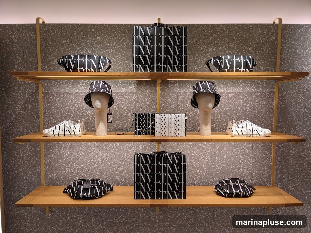 A retail display featuring Valentino branded items, including sneakers, bucket hats, handbags, and clutches, all adorned with the brand's signature VLTN logo pattern in black and white. The items are arranged on wooden shelves against a textured grey wall.