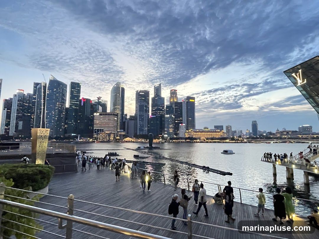 A wide shot of the Marina Bay skyline in Singapore at dusk. The sky is filled with dramatic clouds. In the foreground, a wooden boardwalk is populated with people enjoying the view. The water reflects the city lights. A distinctive building with a prominent Louis Vuitton logo is visible on the right side of the frame.