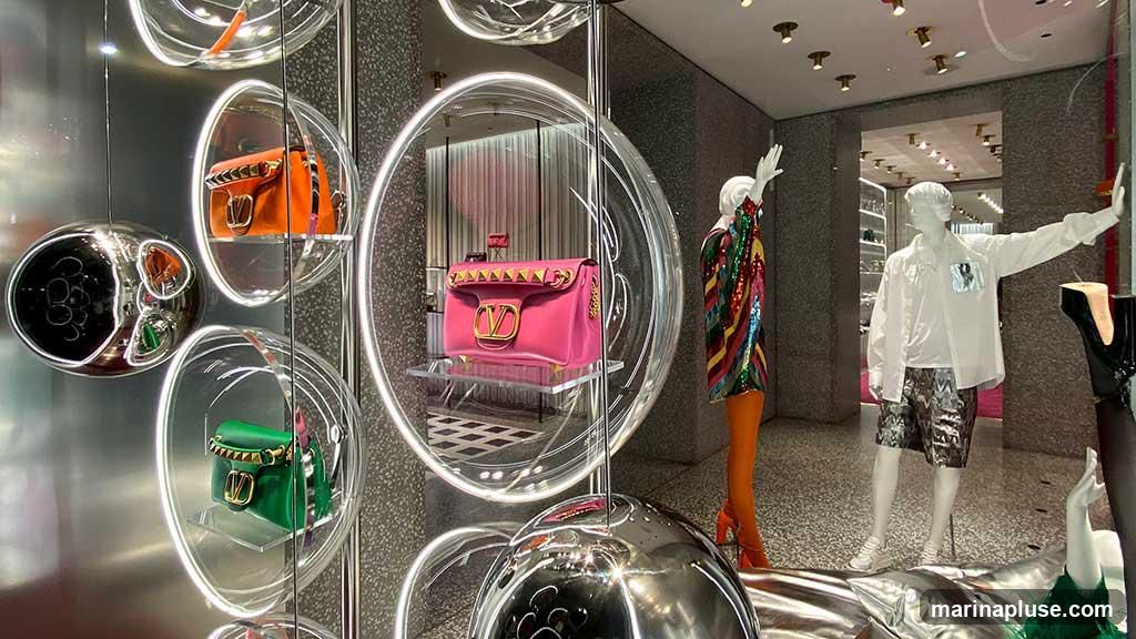 A retail display window featuring several designer handbags in various colors, including pink, orange, and green, showcased within clear, illuminated circular frames. Mannequins dressed in fashionable attire are also part of the display, with one wearing a sequined jacket and orange tights, and another in a white shirt and patterned shorts. The background shows the interior of a high-end store with shelves and lighting.
