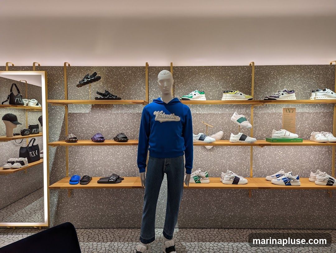 A mannequin wearing a blue hoodie and jeans stands in the center of a shoe store. The mannequin is surrounded by shelves displaying various shoes and bags. The shelves are made of wood and are supported by metal frames. The background wall is textured with a speckled pattern. To the left, a mirror reflects more merchandise.