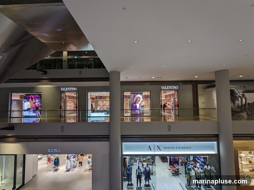 An interior view of a multi-level department store. On the upper level, storefronts for Valentino and Armani Exchange are visible, displaying merchandise and mannequins. On the lower level, a Zara store is seen with clothing racks and displays.