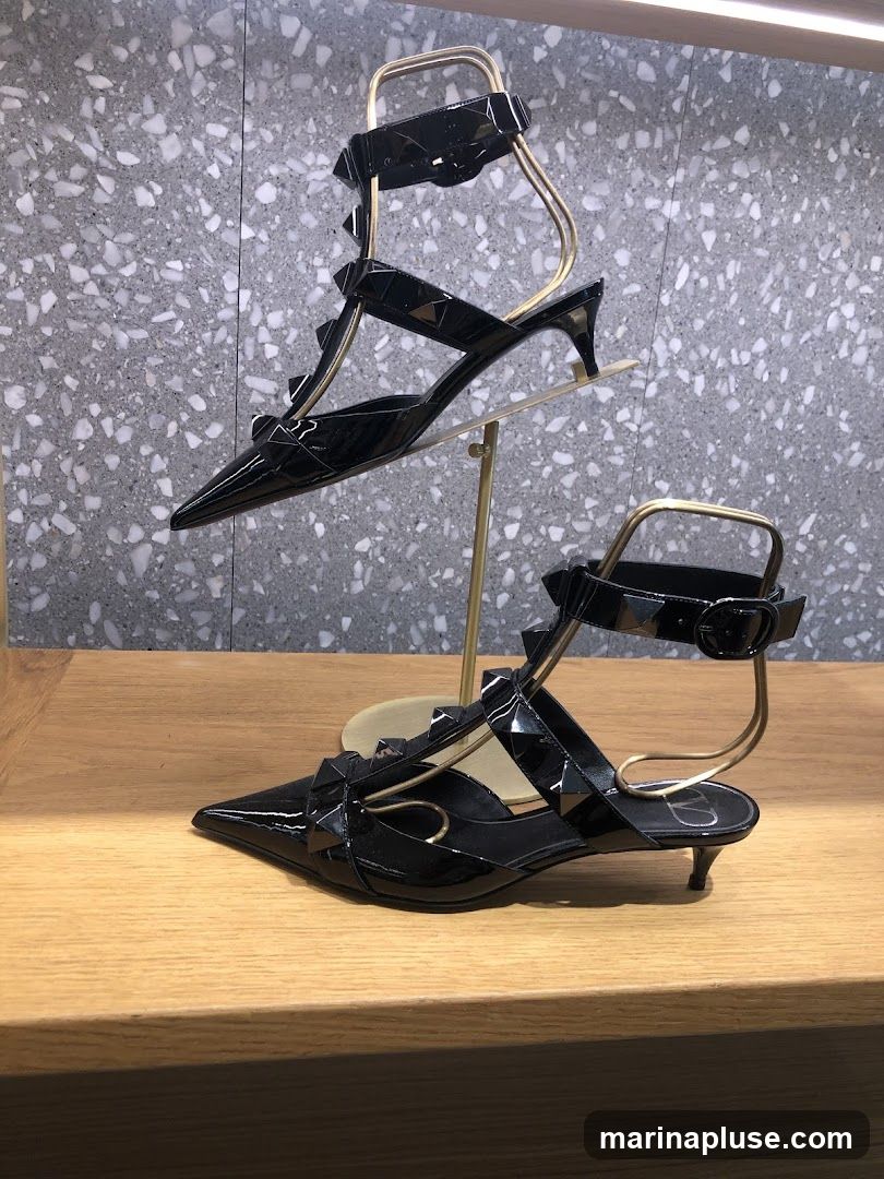 Two pairs of black patent leather heels with ankle straps and pyramid studs are displayed on gold stands against a speckled gray background. The shoes have pointed toes and kitten heels. A small logo is visible on the heel of the lower shoe.