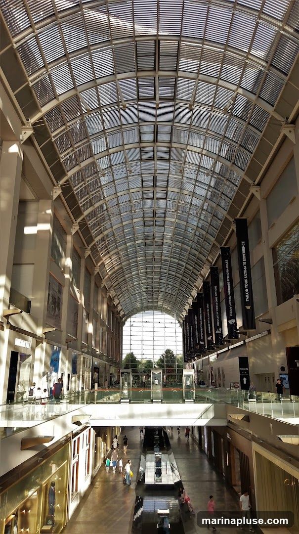 The image shows the interior of a large, modern shopping mall with a high, arched glass ceiling that allows natural light to filter in. The mall has multiple levels with shops and walkways. People are seen walking and browsing. Banners hang from the ceiling, some with text that is partially visible. The architecture is sleek and contemporary, with reflective surfaces on the floor and railings.
