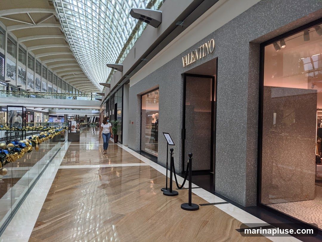 The exterior of a Valentino store in a modern shopping mall. The store has a gray speckled facade with the illuminated "VALENTINO" logo above the entrance. A glass display window is to the right of the entrance. Ropes and stanchions are placed in front of the entrance. The mall interior features a high, arched ceiling with skylights, polished wooden floors, and other storefronts visible in the distance.