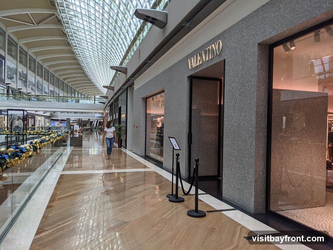 The exterior of a Valentino store in a modern shopping mall. The store has a gray speckled facade with the illuminated "VALENTINO" logo above the entrance. A glass display window is to the right of the entrance. Ropes and stanchions are placed in front of the entrance. The mall interior features a high, arched ceiling with skylights, polished wooden floors, and other storefronts visible in the distance.