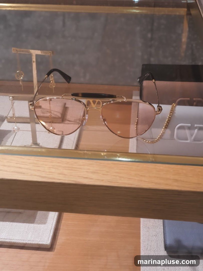 A pair of gold-rimmed sunglasses with pink lenses is displayed on a glass shelf. Behind the sunglasses, a white pouch with a gold logo is visible, along with a gold chain necklace. To the left, earrings are displayed on stands. The display case has a wooden frame.