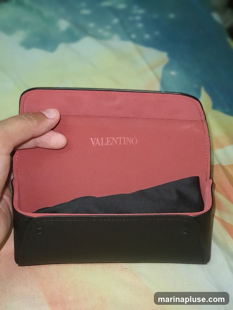 A hand holds open a black and red Valentino branded eyewear case. The interior is lined with red fabric and embossed with the Valentino logo. A black cloth is visible inside the case.