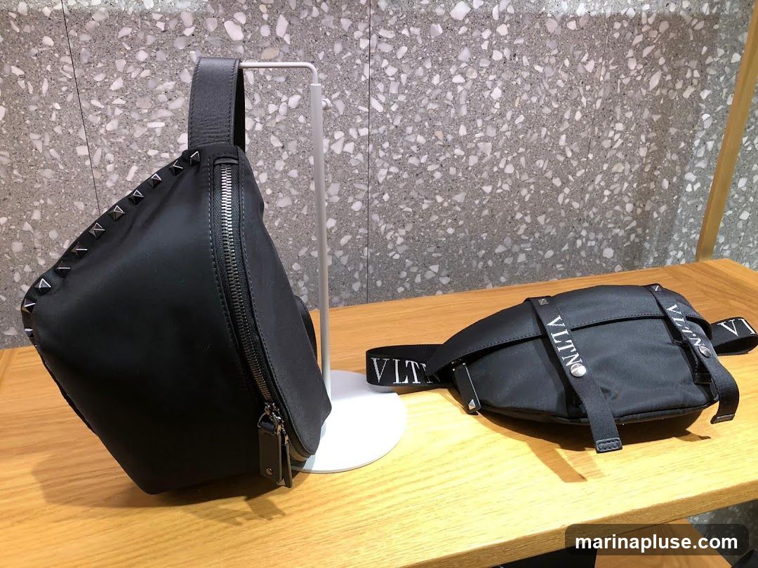 Two black bags are displayed on a wooden surface. The bag on the left is a sling bag with pyramid studs along the top edge and a zipper closure. It is displayed on a white stand. The bag on the right is a fanny pack with "VLTN" printed on the strap.