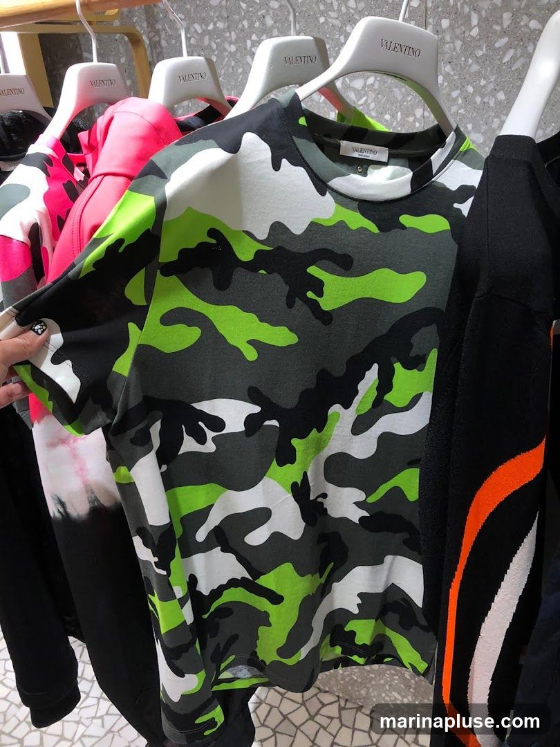 A close-up shot of a camouflage t-shirt with neon green, white, and dark gray patterns. The t-shirt is hanging on a rack with other clothing items, including a pink hoodie and a black garment with orange and white stripes. The brand name "VALENTINO" is visible on the hangers.