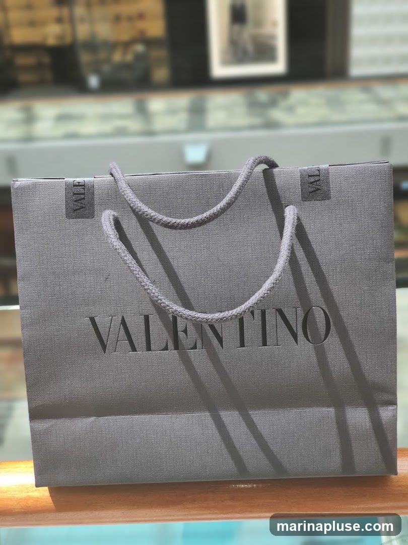 A gray Valentino shopping bag with braided handles sits on a wooden surface. The Valentino logo is embossed on the front of the bag. The background is blurred, showing what appears to be a retail environment with displays and artwork.