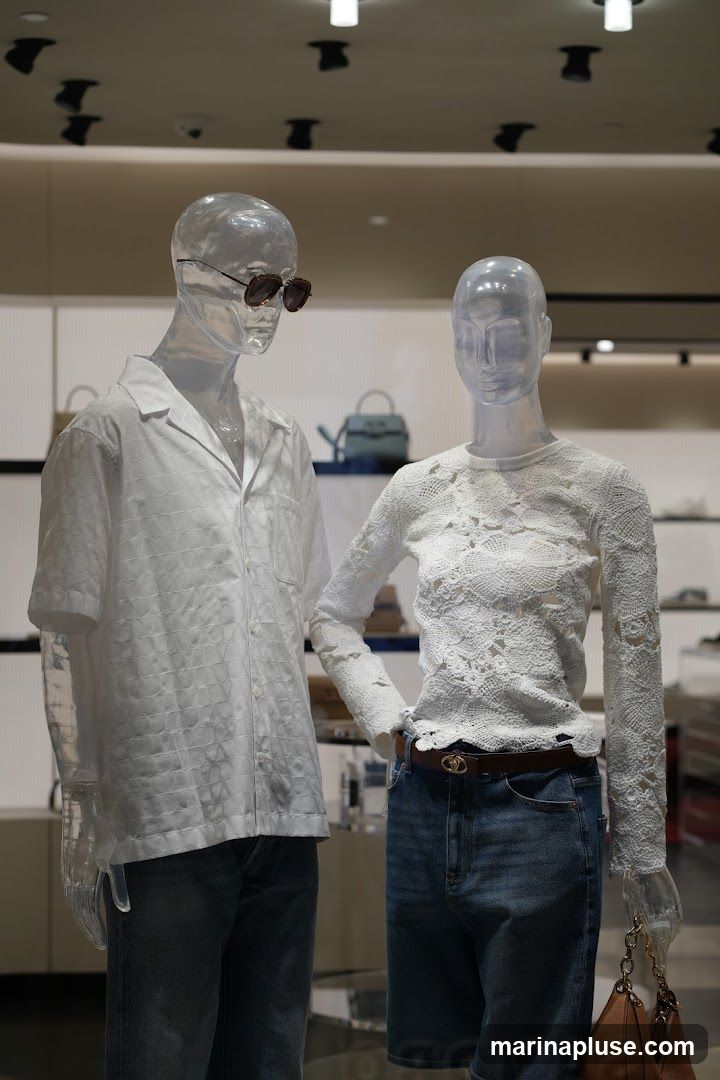 Two clear mannequins are displayed in a store. The male mannequin on the left wears a white short-sleeved patterned shirt and jeans. He has sunglasses on. The female mannequin on the right wears a white lace long-sleeved top and denim shorts, accessorized with a brown belt and holding a brown handbag. In the background, shelves display handbags and other merchandise.