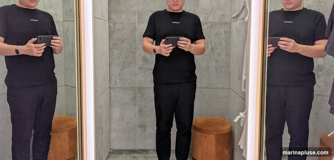 A person stands in a fitting room, wearing a black t-shirt and black pants. The t-shirt has a small white logo on the chest. The person is holding a smartphone in both hands. The fitting room has mirrors on the sides and a grey tiled wall. There are two brown ottomans visible in the reflections. A white hanger is visible on the right side of the image.