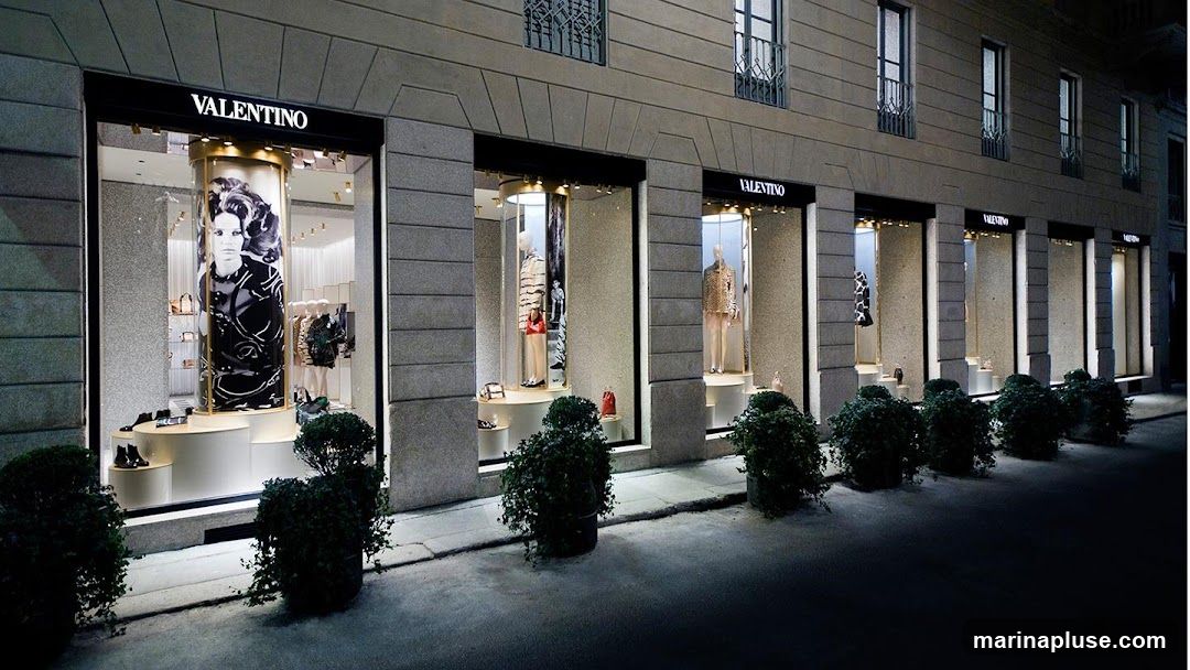 A nighttime exterior view of a Valentino store with multiple display windows showcasing mannequins dressed in clothing and accessories. The store's name, VALENTINO, is prominently displayed in illuminated lettering above each window. The windows feature elegant displays with spotlights highlighting the merchandise. Green bushes are planted in front of the store.
