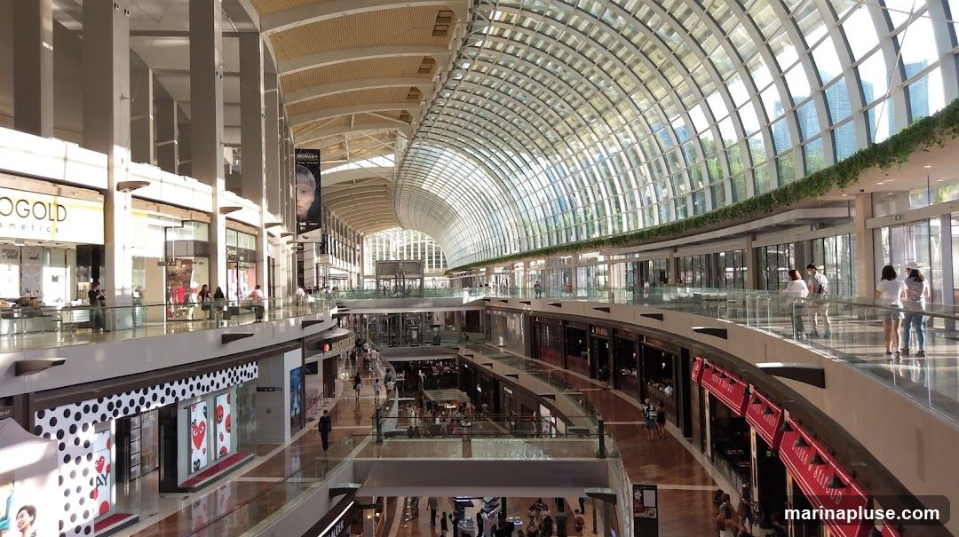 A wide-angle view of the interior of a large, modern shopping mall. The architecture features a curved glass ceiling that allows natural light to flood the space. Multiple levels of shops and walkways are visible, with shoppers browsing and walking around. Some shops have prominent signage, including one with "GOLD COSMETICS" and another with a black and white polka dot pattern. Banners hang from the ceiling, and greenery is incorporated into the upper levels.