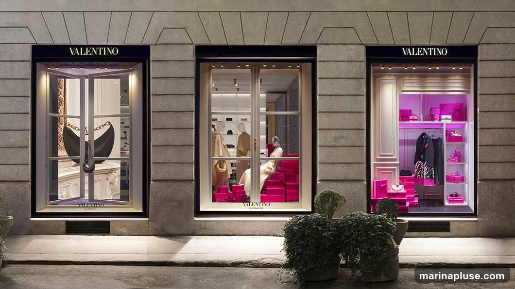 The exterior of a Valentino store at night, featuring three large display windows. The left window showcases a black handbag with a gold chain. The middle window displays mannequins dressed in neutral-toned clothing, surrounded by pink gift boxes. The right window is illuminated with pink light and displays clothing, handbags, and shoes, also with pink gift boxes. The Valentino logo is visible above each window.