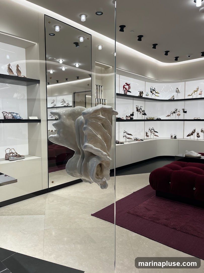 View through a glass door into a brightly lit, modern shoe store. Shelves display a variety of high-heeled shoes and sandals. A plush, dark red sofa is visible on the right, with a matching rug. The door handle is an ornate, sculptural piece.
