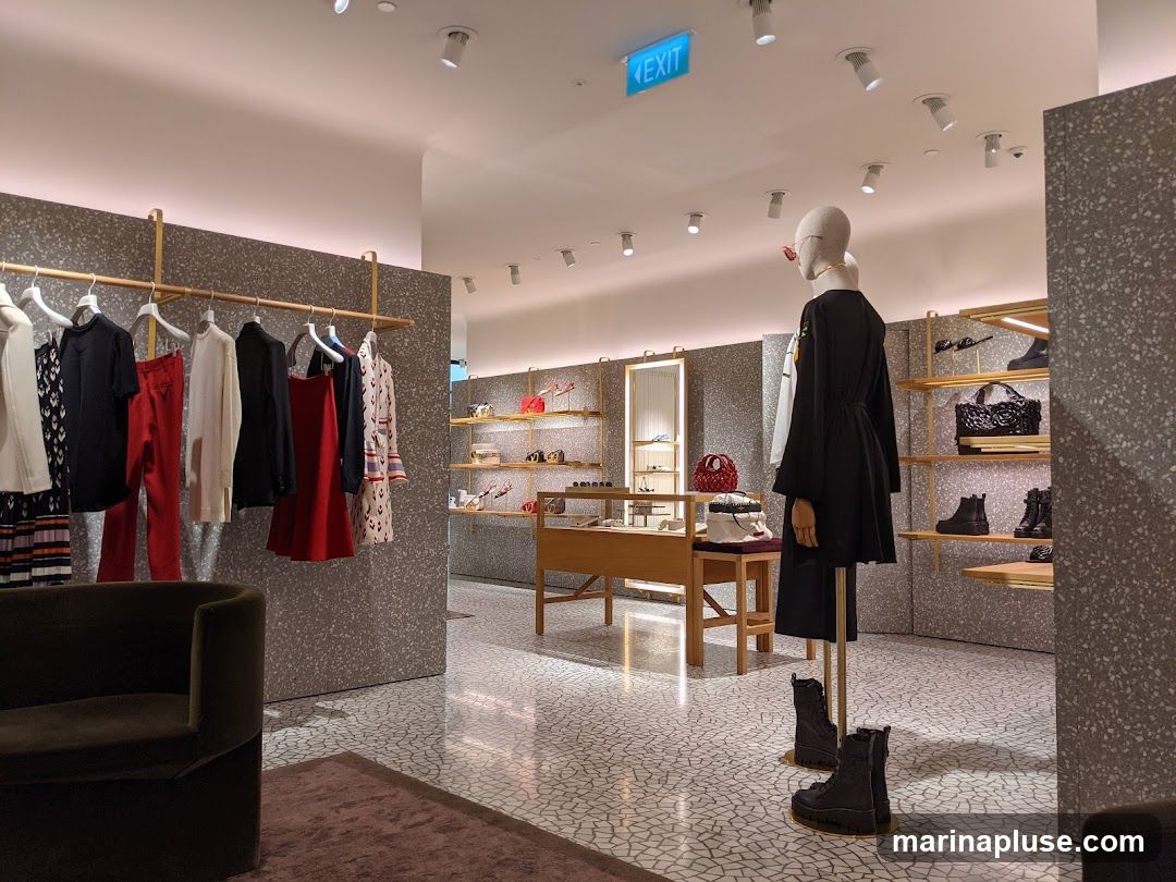 A well-lit fashion boutique interior with clothing displayed on racks and shelves. A mannequin stands in the foreground wearing a black dress and boots. Various handbags and shoes are visible on display shelves in the background. The store features a modern design with speckled walls and a patterned floor.