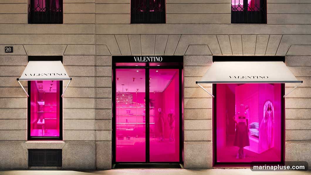 The exterior of a Valentino store is shown at night. The facade is made of stone. There are three large windows and a central double door. Each window and the door are illuminated with bright pink light, creating a vibrant display. White awnings with the Valentino logo are above the windows and the door. The windows display mannequins dressed in clothing and accessories. The central doors also reveal a pink-lit interior with displays and chandeliers.