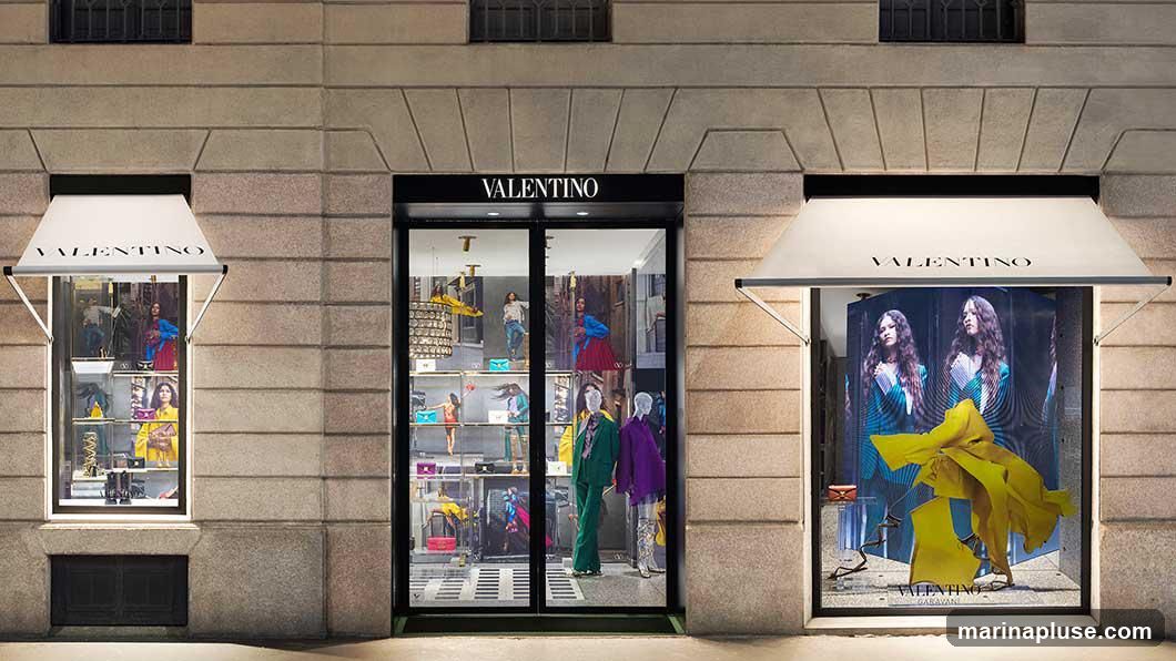 The exterior of a Valentino store at night. The facade features large display windows showcasing mannequins dressed in clothing and accessories. Above the windows, awnings with the Valentino logo are visible. The main entrance is a glass door with the Valentino logo above it.