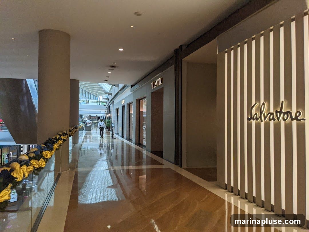 A well-lit interior hallway of a modern shopping mall with polished floors reflecting the lights. On the right, the storefront of Salvatore Ferragamo is visible, featuring its name in a stylized script illuminated by soft lighting. The facade is decorated with vertical white slats. To the left, the storefront of Valentino is also visible, with its name displayed above the entrance. A security guard is walking down the hallway in the distance.