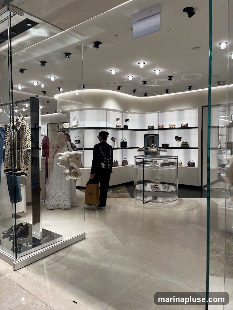 A view into a brightly lit, modern luxury retail store. The interior features sleek white shelving displaying various handbags and accessories. In the foreground, a person is seen holding a shopping bag with a visible logo. To the left, a display case shows clothing and shoes. The overall ambiance is clean, elegant, and upscale.