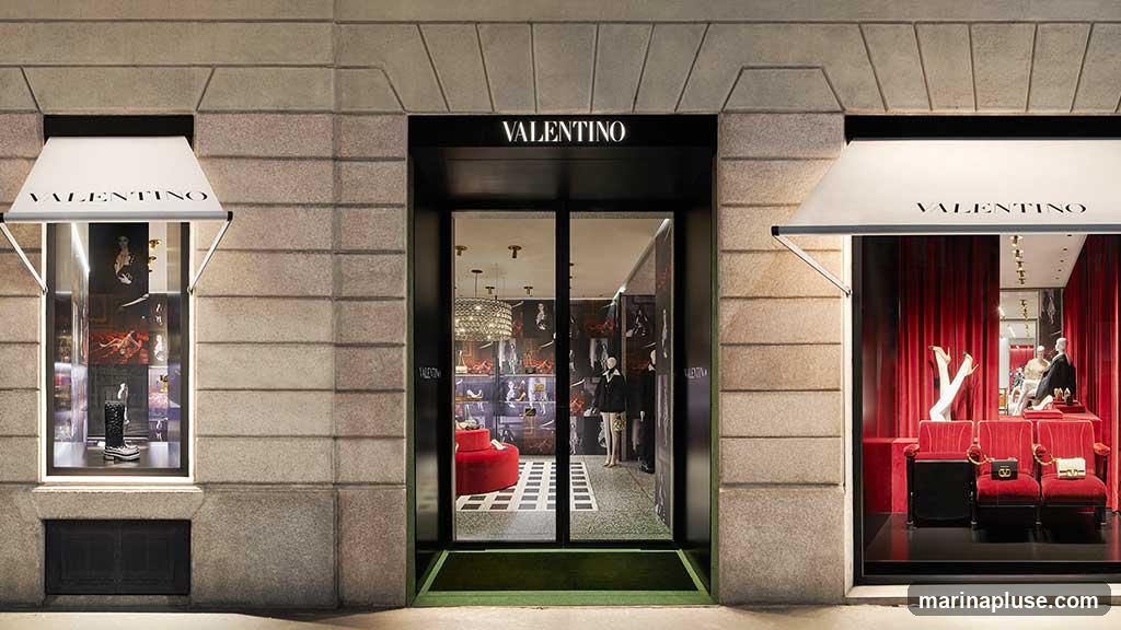 The exterior of a Valentino store at night. The entrance is a double glass door with a black frame. Above the entrance, the word "VALENTINO" is illuminated in white letters. To the left and right of the entrance are display windows with awnings above them, also displaying the "VALENTINO" logo. The windows showcase mannequins and merchandise, with red velvet curtains visible in the right window. The building facade is made of stone blocks.