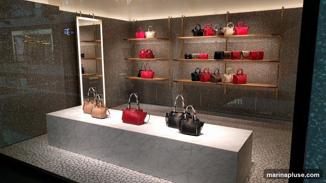 A retail display window showcasing a collection of designer handbags on shelves and a marble pedestal. The handbags are in various colors including red, black, white, and beige, and feature stud embellishments. The store interior has a speckled grey wall and wooden shelves.
