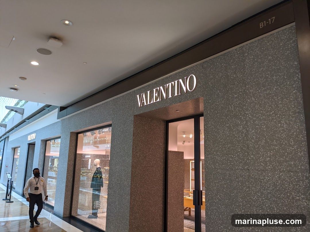 The exterior of a Valentino store with its name illuminated above the entrance. The store features large display windows showcasing mannequins and merchandise. A person in a white shirt and black pants, wearing a mask, walks past the storefront. The store is located in a mall, indicated by the ceiling and lighting.
