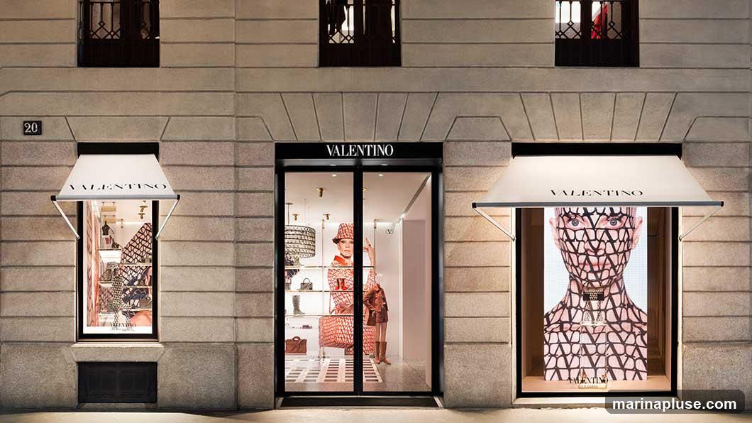 The exterior of a Valentino store at night. The facade is made of stone blocks. There are three large display windows and a central entrance. The name VALENTINO is displayed above the entrance and on awnings over two of the windows. The windows display mannequins and merchandise. One window features a large digital display of a face covered in the Valentino V logo pattern.