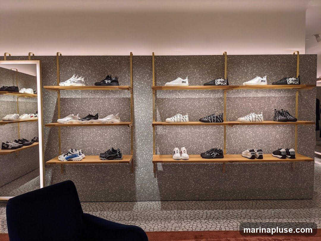 A well-lit interior of a modern shoe store. Shelves are stocked with various sneakers, displayed in pairs. The shelves are made of wood and supported by gold-colored metal frames. The background wall has a speckled texture. A large mirror is visible on the left, reflecting more shelves and shoes. A dark blue armchair is in the foreground.