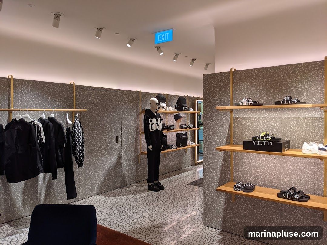 A well-lit interior of a modern fashion boutique. Racks of black clothing hang on the left, with a mannequin dressed in a black hoodie and pants standing in the center. Shelves on the right display various footwear, including sandals and sneakers, with some items featuring the "VLTN" logo. An "EXIT" sign is visible on the ceiling.