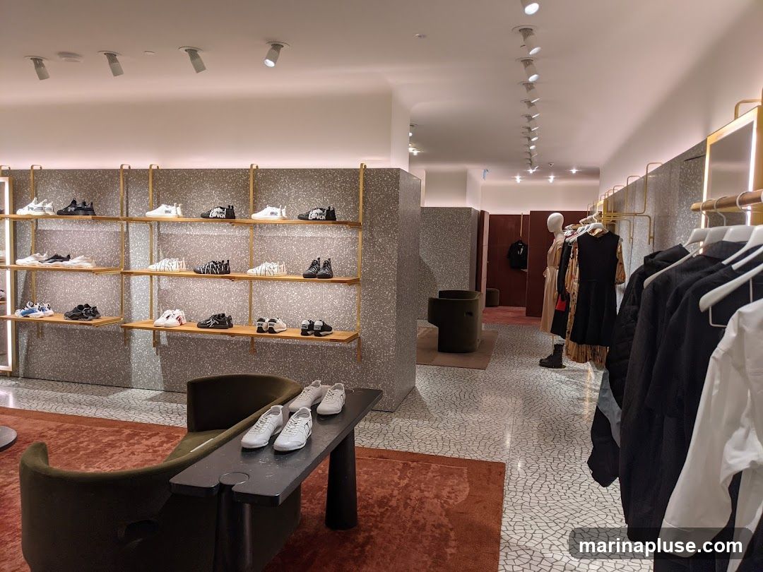 The interior of a modern retail store, featuring shelves stocked with various sneakers and racks of clothing. A display table in the foreground shows several pairs of white sneakers. Mannequins are visible in the background, dressed in apparel. The store has a clean, minimalist aesthetic with terrazzo-patterned walls and floors.