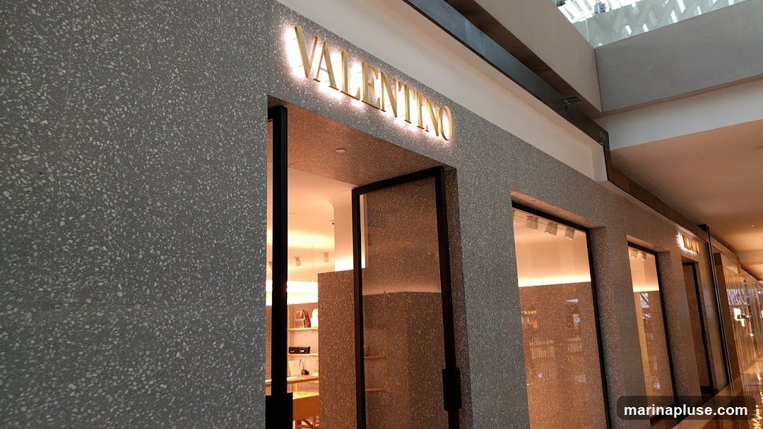 The exterior of a Valentino store with a terrazzo-like facade. The store name "VALENTINO" is displayed in illuminated gold lettering above the entrance. The entrance is open, revealing a brightly lit interior with displays of merchandise. Several display windows are visible along the facade.