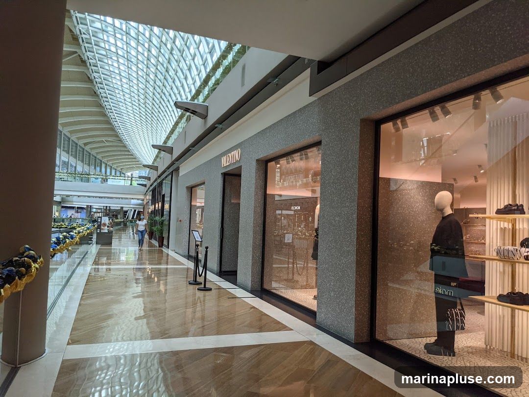 A Valentino store with a large display window showcasing a mannequin dressed in black, a handbag, and shoes. The store has a speckled gray facade and the Valentino logo is visible above the entrance. The interior of the store is visible through the window, with shelves displaying merchandise. The storefront is located in a well-lit indoor shopping mall with a distinctive arched ceiling.