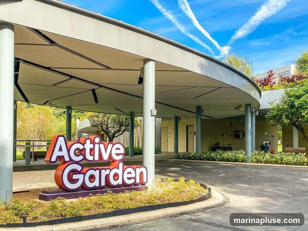 A large, three-dimensional sign spelling out "Active Garden" is prominently displayed in front of a modern, covered entrance. The sign has orange and white lettering with a purple base. The entrance features a curved, white roof supported by grey pillars. Beyond the entrance, there are doors and some outdoor amenities visible. The sky above is blue with white contrails.