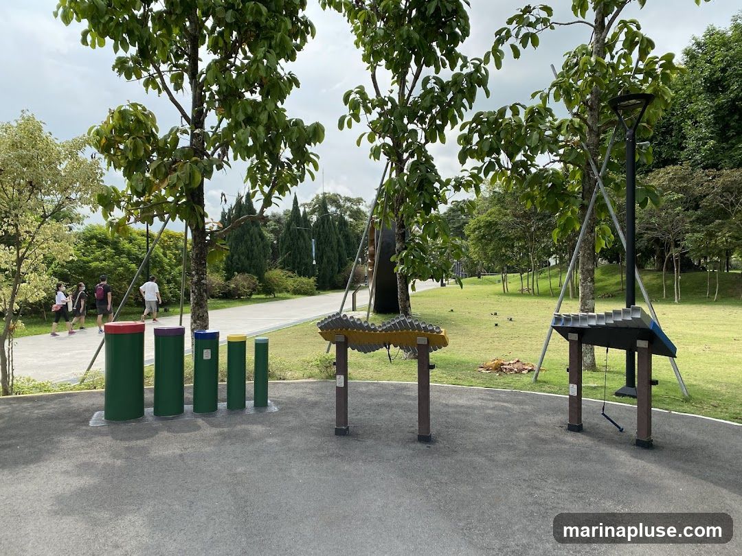 A park setting with various outdoor musical instruments. In the foreground, there are several large, colorful cylindrical objects that appear to be part of a musical installation. To the right, two larger structures with metal bars are set up, resembling xylophones or glockenspiels. People are walking on a paved path in the background, surrounded by lush green trees and grass. The sky is overcast.