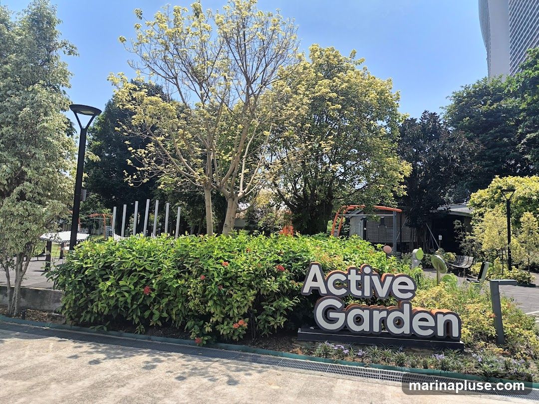 A sign that reads "Active Garden" is displayed in front of lush green bushes and trees. The sign has large, three-dimensional letters with a white front and a gray outline. Behind the sign, there are various plants, trees, and some structures, including a small building and playground equipment. The sky is clear and blue.