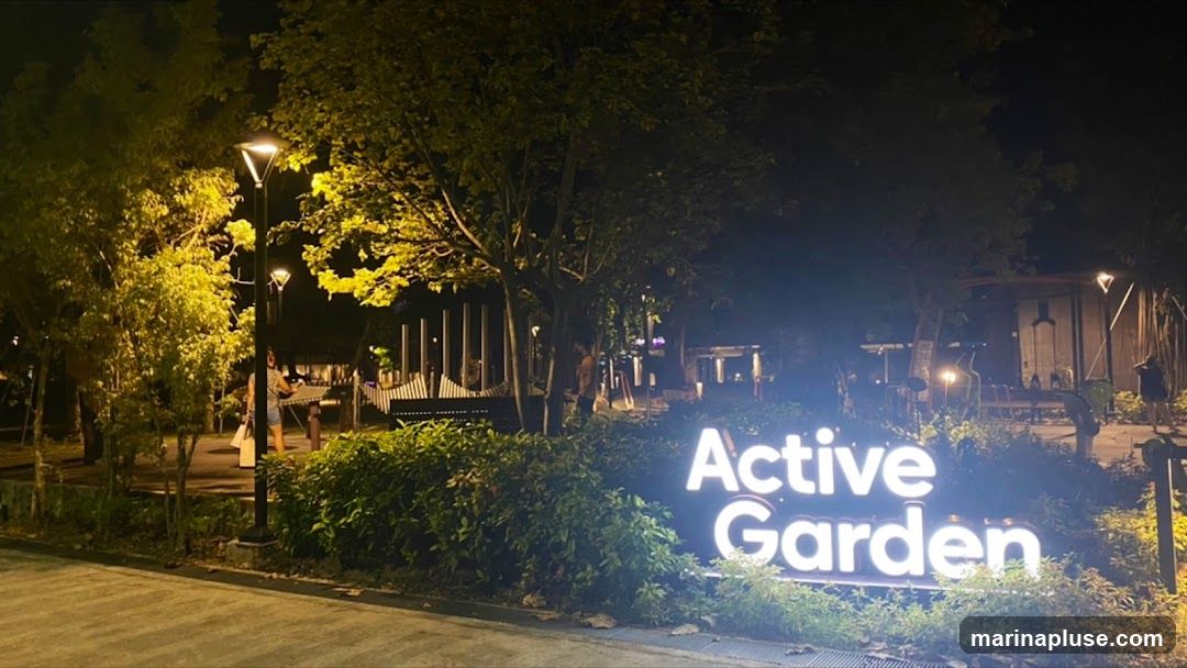 A large, illuminated sign spelling out "Active Garden" is prominently displayed at night, surrounded by lush greenery and trees. The sign is lit with bright white lights, making it stand out against the dark background. Several lampposts illuminate the surrounding park area, casting a warm glow on the trees and pathways. In the background, there are glimpses of park structures and people enjoying the outdoor space.