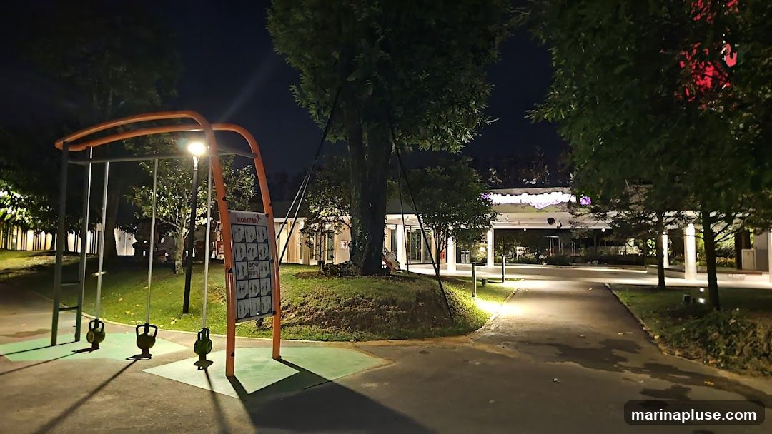 An outdoor recreation area is shown at night. In the foreground, there is a playground structure with swings and a sign displaying exercise instructions. A paved path leads towards a building in the background, which appears to be a commercial establishment with illuminated signage and glass doors. Trees and landscaping are visible throughout the scene, with artificial lighting illuminating the area.
