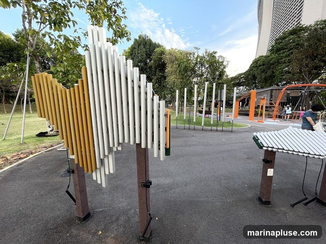 An outdoor park area with various musical instruments, including a large chime-like structure and a xylophone-like instrument. In the background, there is a playground structure and a tall modern building. Several people are visible in the background near the playground.