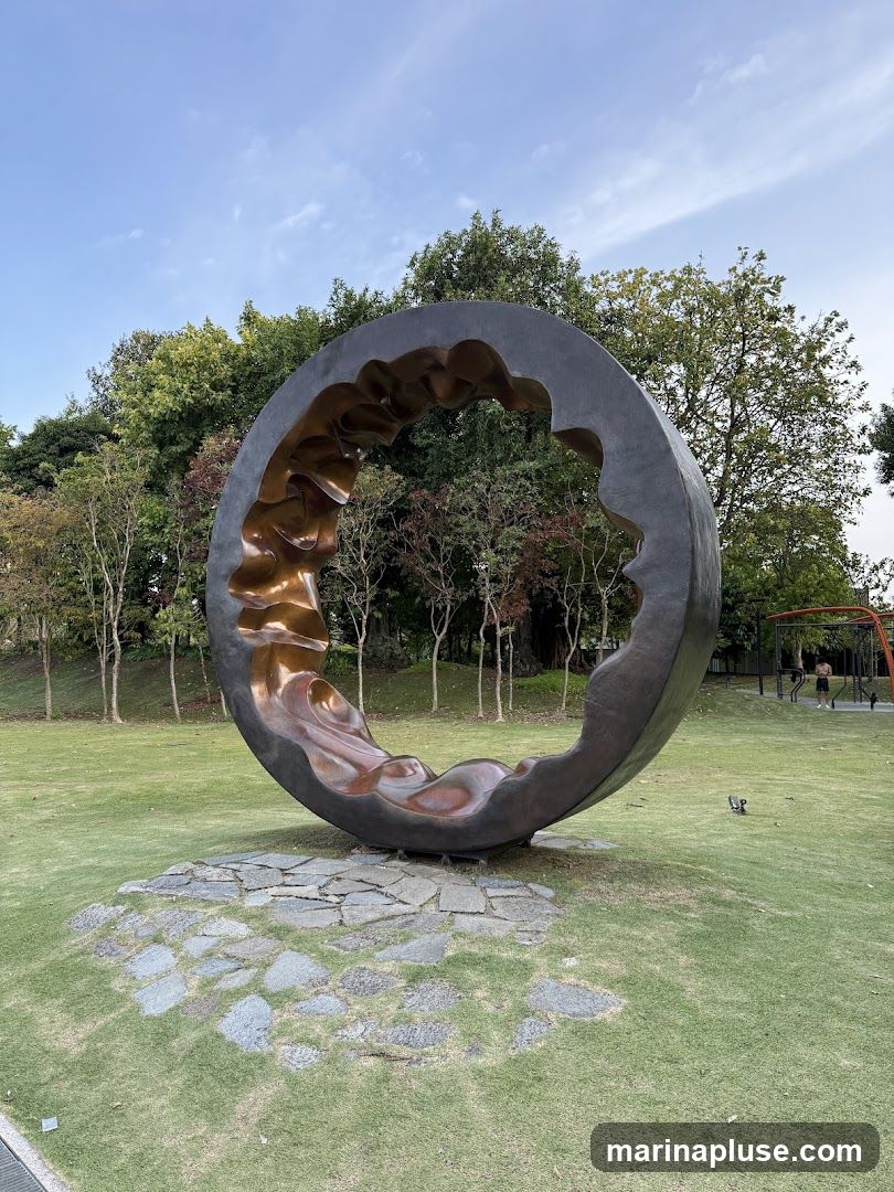 A large, circular sculpture with a bronze-colored, wavy interior and a dark exterior is displayed on a grassy lawn. The sculpture is surrounded by a base of irregular stone slabs. In the background, there are trees with green and some reddish foliage, and a clear blue sky with scattered clouds. To the right, a person is visible near some outdoor exercise equipment.