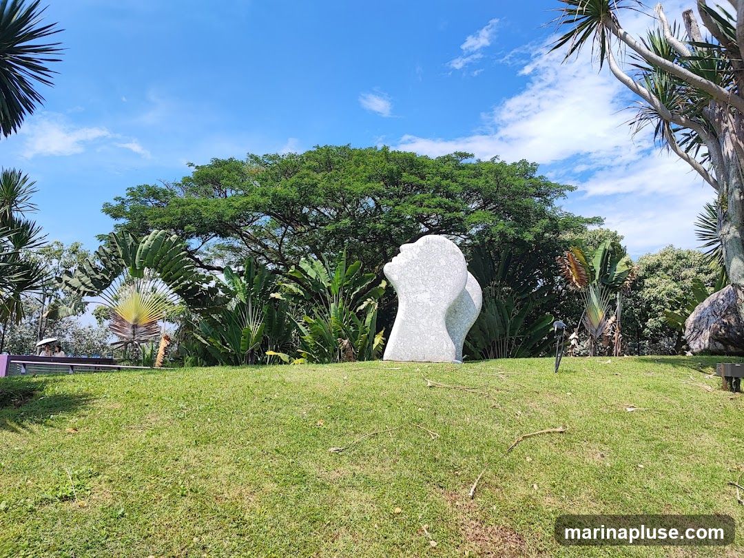 A white abstract sculpture resembling two heads is situated on a grassy hill in a park. Lush green trees and tropical plants surround the sculpture, with a bright blue sky and scattered clouds overhead.