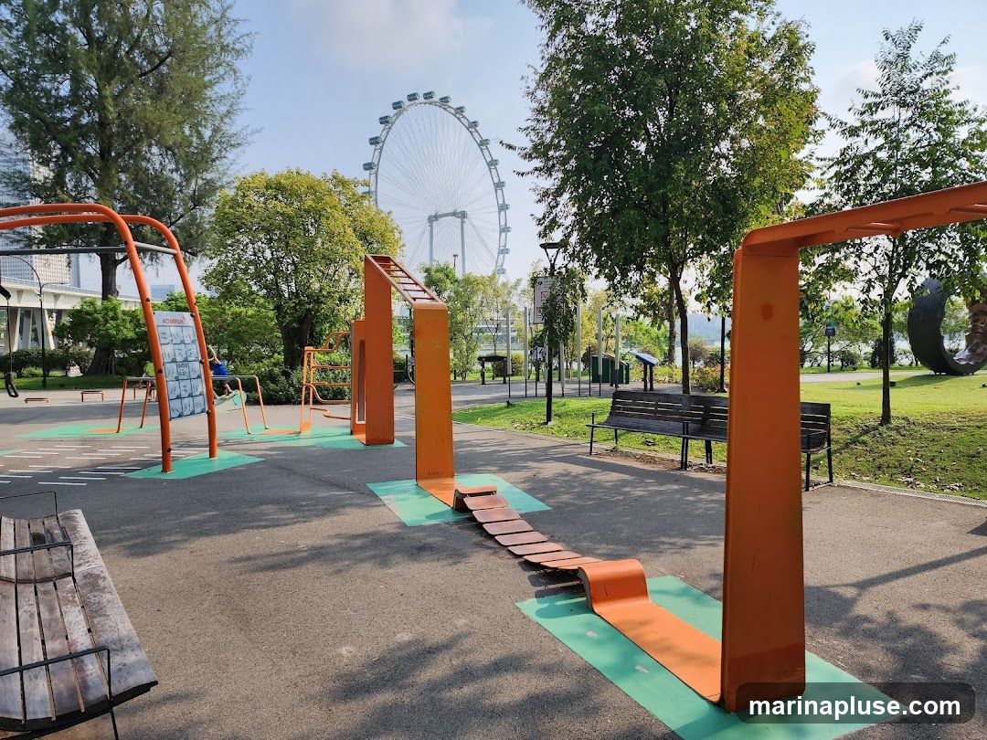 An outdoor fitness park with various exercise equipment, including monkey bars, balance beams, and pull-up bars. In the background, a Ferris wheel and trees are visible under a clear sky.