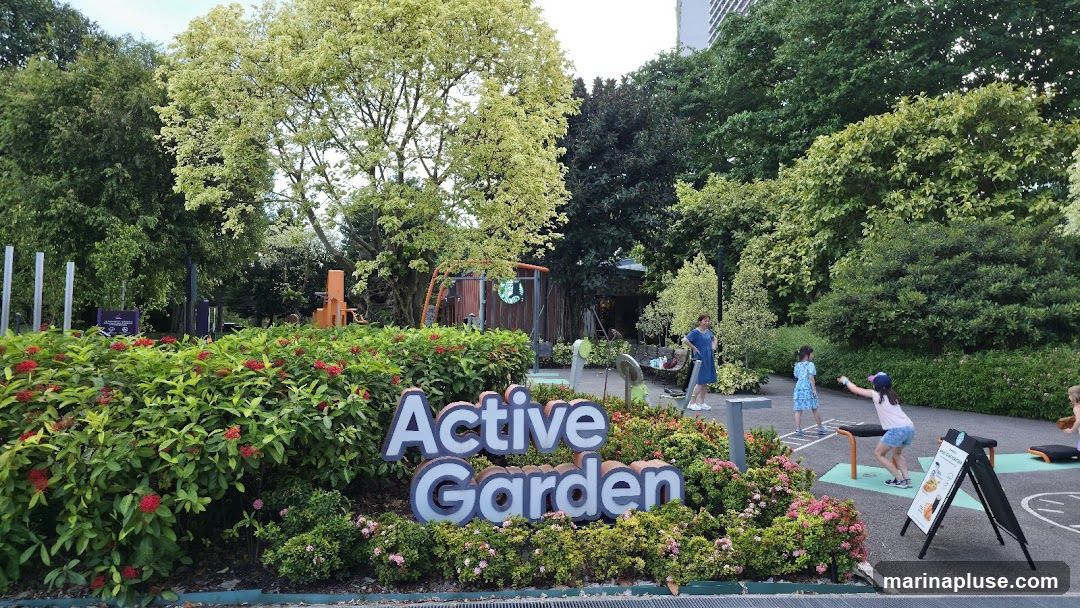 A park scene with lush greenery and flowering bushes. In the foreground, a large sign reads "Active Garden". Several children are playing on exercise equipment in the background. A Starbucks logo is visible on a small structure.