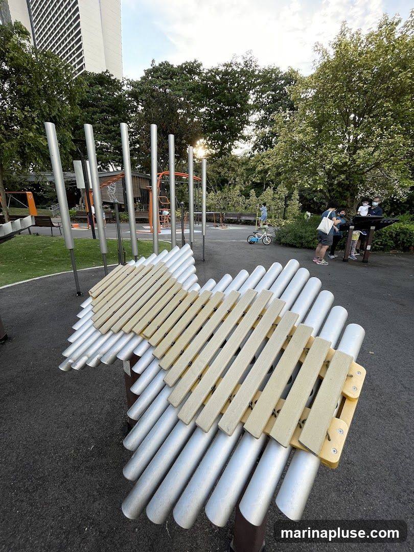 A playground with large outdoor musical instruments, resembling xylophones, made of metal tubes and wooden bars. Several people are present, including children and adults, interacting with the instruments and playground equipment. The scene is set in a park-like environment with trees and a tall building in the background.