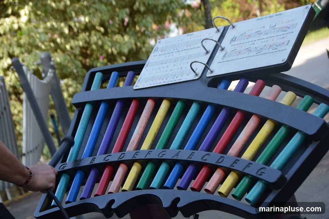 A colorful outdoor musical instrument, resembling a xylophone or glockenspiel, with metal bars of various colors. A songbook with sheet music for "Bingo" and "Rock-a-bye Baby" is attached to the instrument. The instrument is set up outdoors, with blurred greenery in the background. A hand is visible on the left, holding a mallet, suggesting the instrument is in use.